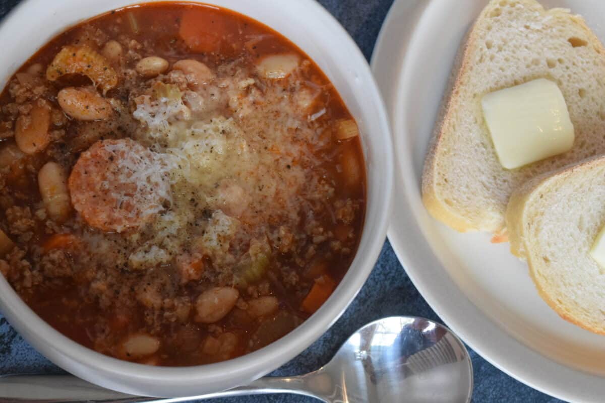 a white bowl of pasta e fagioli soup with a plate of buttered french bread and a soup spoon