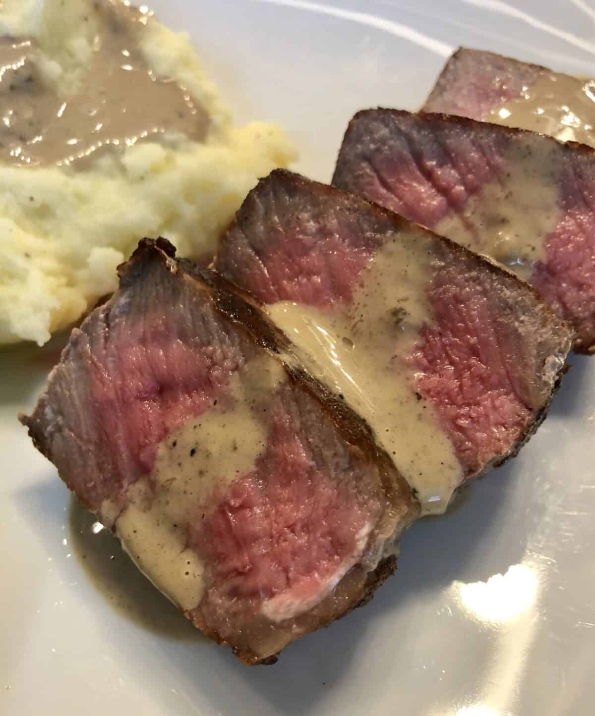 White plate of sliced steak with mashed potatoes and drizzled with cognac cream sauce