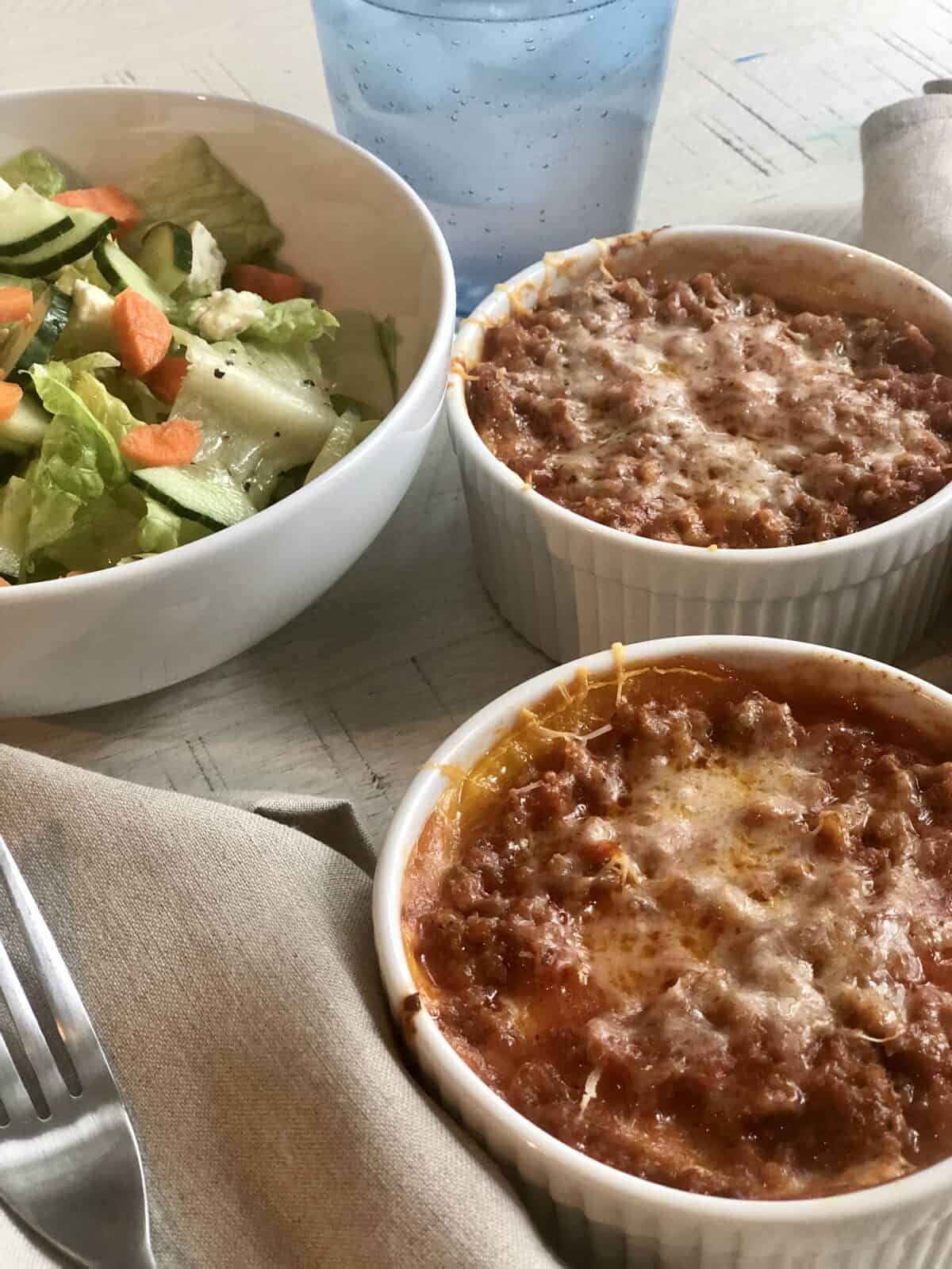 two individual butternut squash lasagnas in white ramickens with a tan napkin fork and a bowl of salad a tan napkin and a glass of lemonade