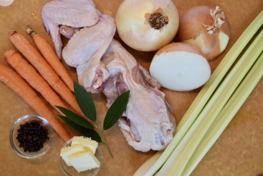 a wood cutting board with homemade chicken stock ingredients including black peppercorns in a  glass bowl 4 carrots 3 bay leaves butter a chicken back and wings 2 onions and 3 celery stalks