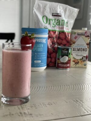 low sugar strawberry smoothie in a glass and garnished with a strawberry and collagen protein frozen strawberries almond milk and coconut cream in the back round