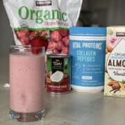 a low sugar strawberry smoothie in a glass with a strawberry for garnish in front of smoothie ingredients including frozen strawberries canned coconut cream collagen peptides and kirkland signature almond milk