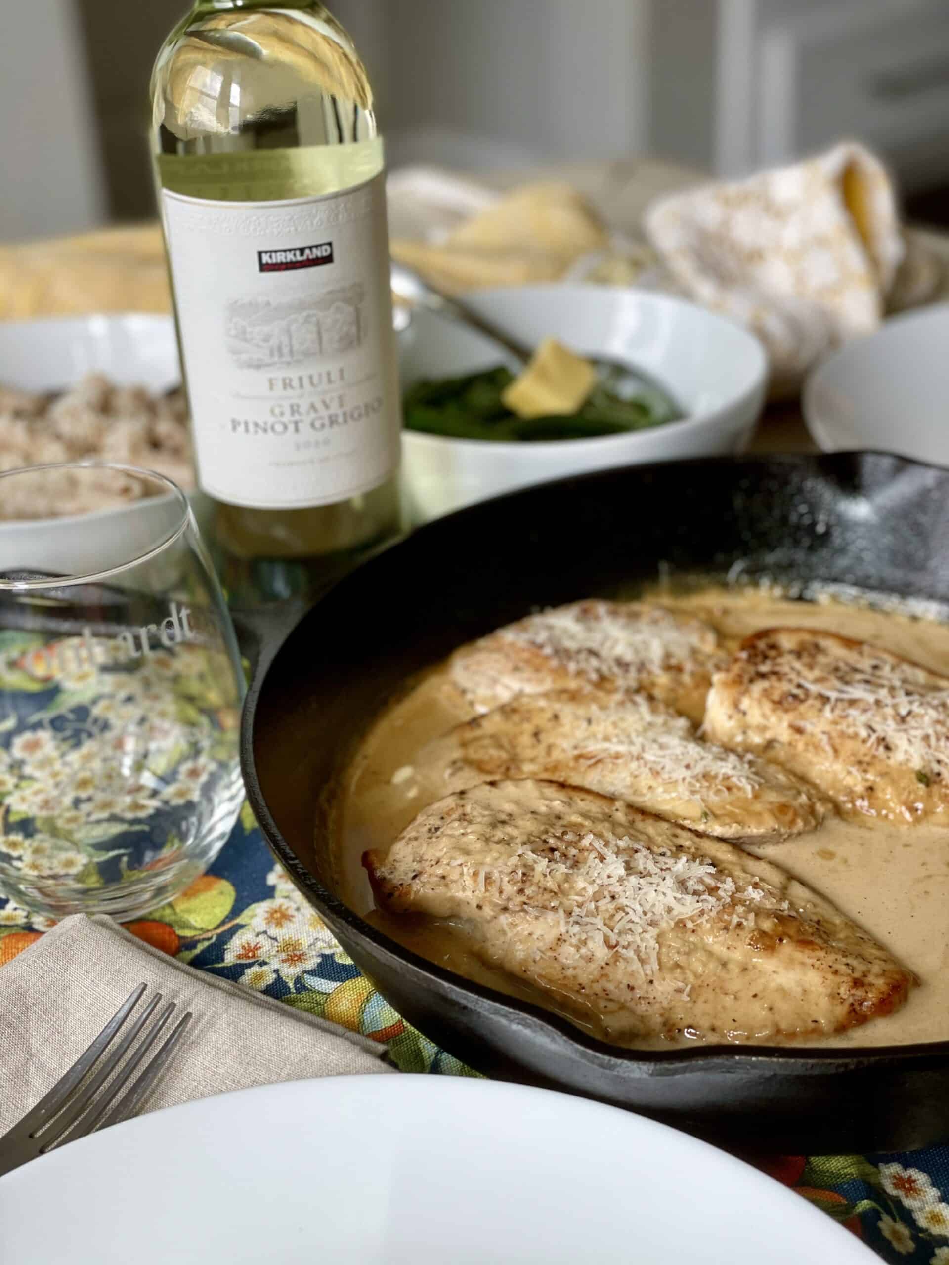 cast iron skillet with creamy lemon chicken set on a table with white plates wine glasses and a bottle of pinot grigio wine
