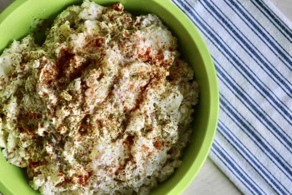 a green bowl of prepared potato salad garnished with paprika and dried parsley set next to a blue and white tea towel