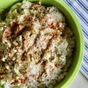 a green bowl of prepared potato salad garnished with paprika and dried parsley set next to a blue and white tea towel