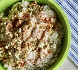 a green bowl of prepared potato salad garnished with paprika and dried parsley set next to a blue and white tea towel