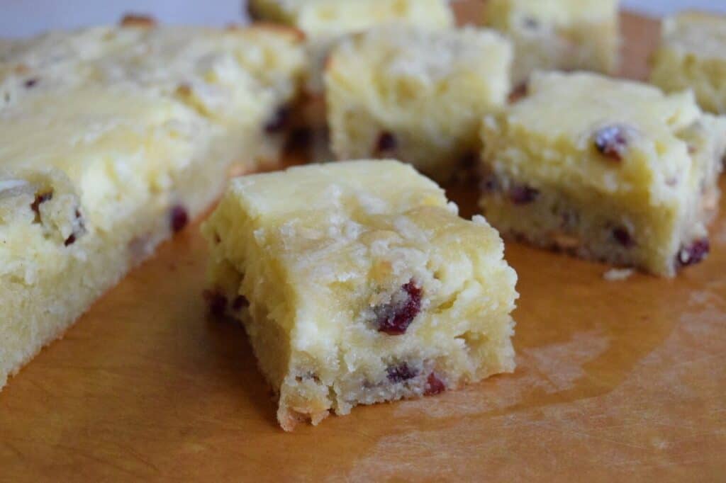 A blondie with a cheesecake filling, white chocolate chips, and dried cranberries on a cutting board.