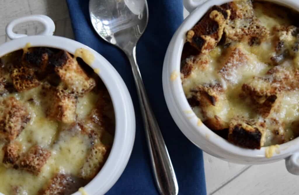 Two white bowls of french onion soup with a blue napkin and soup spoon