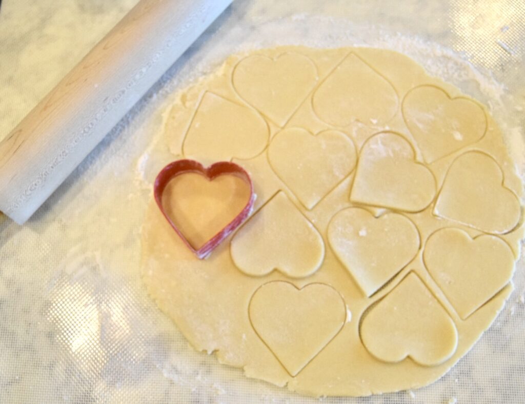 sugar cookie dough rolled out with heart shapes cut into it and a heart shaped cookie cutter still on the board with a rolling pin