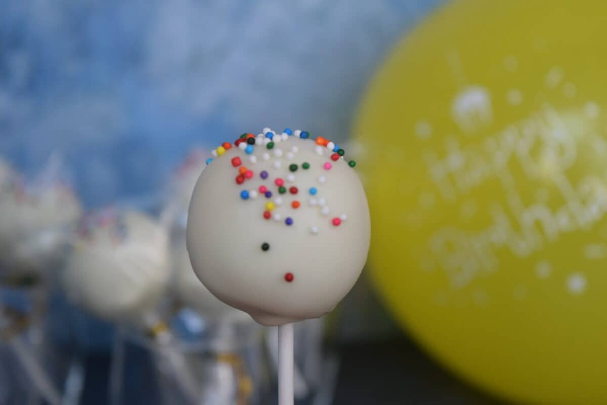 white cake pop with rainbow sprinkles, yellow balloon, and blue background.