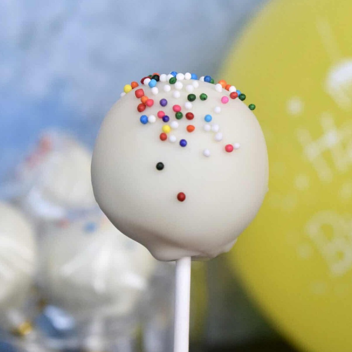 white cake pop with rainbow sprinkles and more cake pops and a yellow balloon in the background