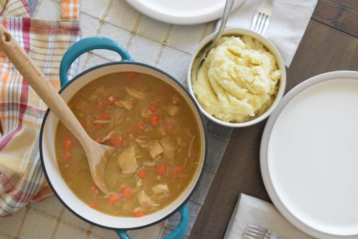 dining table with a teal pot of rotisserie chicken stew a bowl of mashed potatoes 2 plates forks napkins and an apron