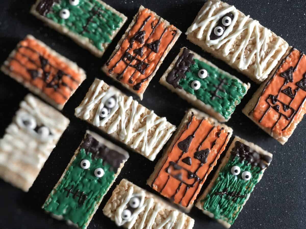 12 rice krispie treats laying flat on black cutting board decorated to look like jack-o-lanterns mummies and frankenstein