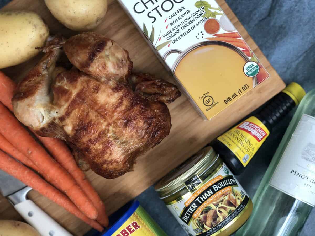 Cutting board with rotisserie chicken box of kirkland signature chicken stock 4 whole carrots a knife organic better then bouillon pinot grigio wine bottle 2 yellow potatoes kitchen bouquet and a blue back drop