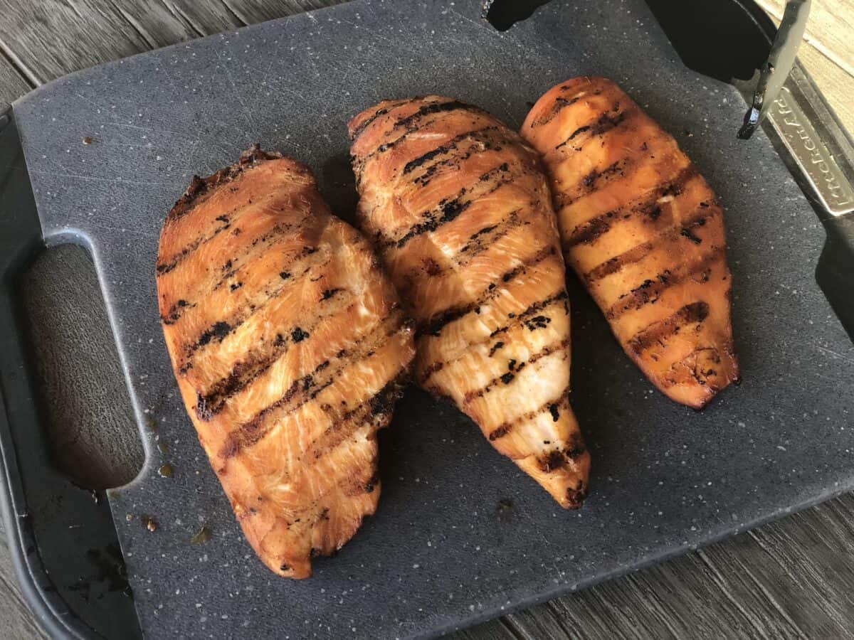 grey cutting board with three grilled pieces of chicken cooked with a freezer friendly chicken marinade and a set of tongs on a wood table