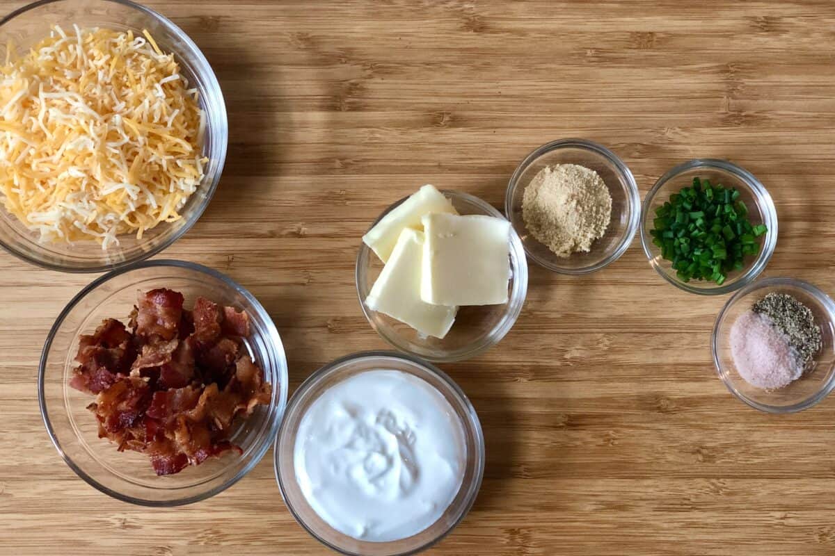 wood cutting board with glass bowls of ingredients including shredded cheese bacon sour cream butter onion powder chives salt and pepper