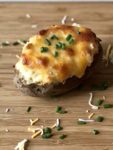 twice baked potato covered in melted cheese and chives on a wood cutting board with cheese and chives sprinkled around it