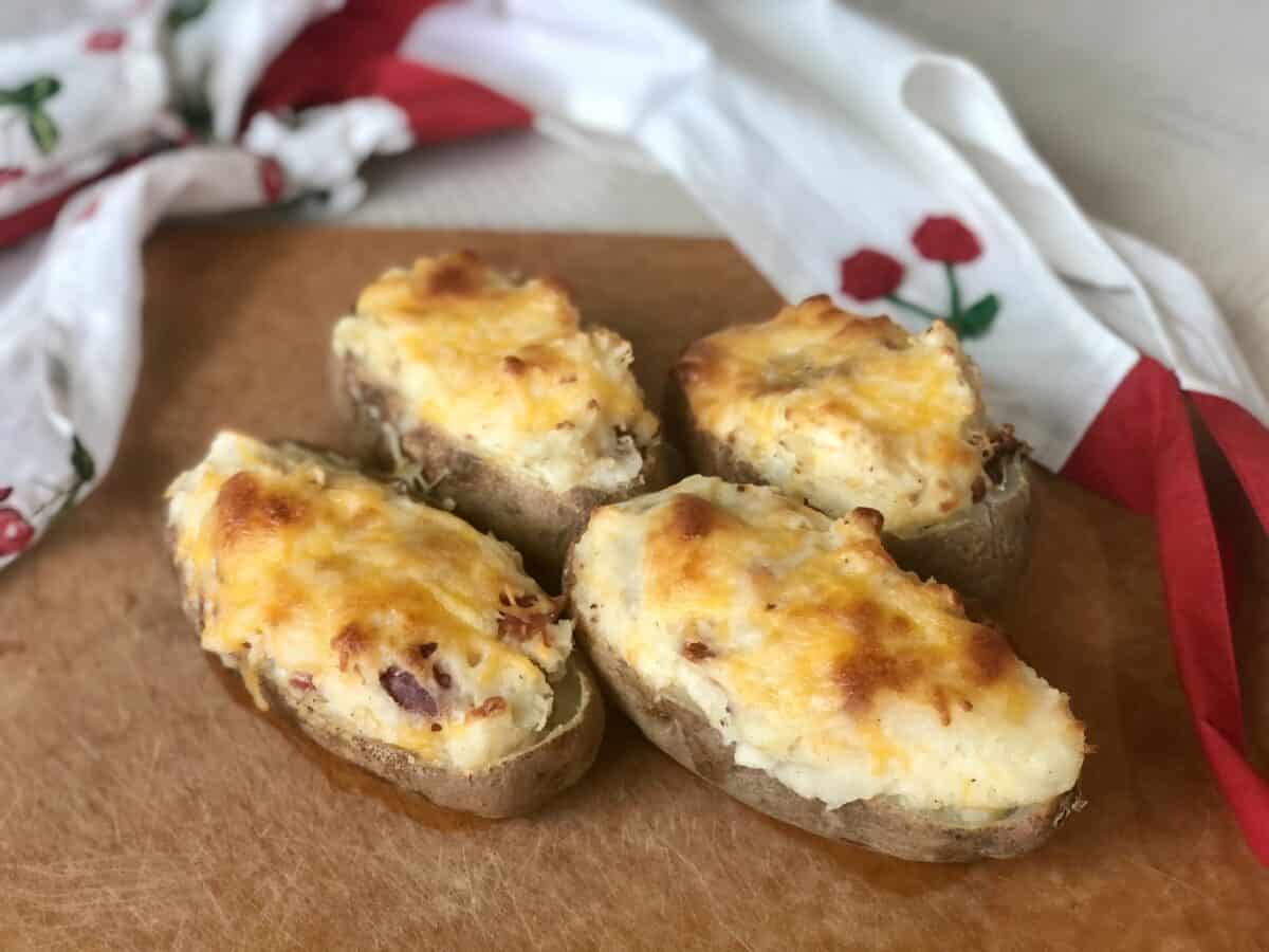 a brown cutting board with 4 easy twice baked potatoes topped with melted cheese and an apron in the backround