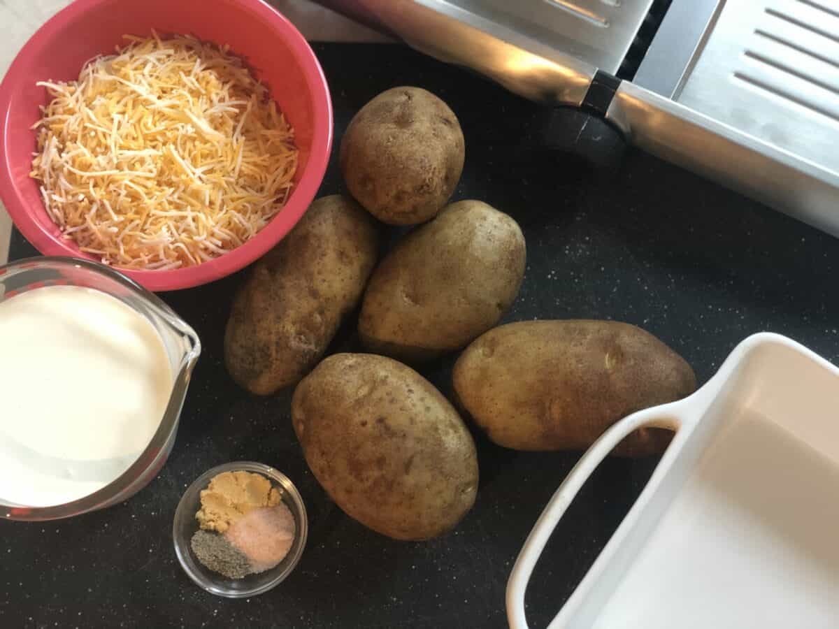 5 russet potatoes on a black cutting board next to a measuring cup of cream red bowl of shredded cheese a small bowl of spices with a partial view of a mandolin and white baking dish