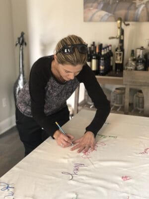 A woman signing a tablecloth that is spread out on a dining room table