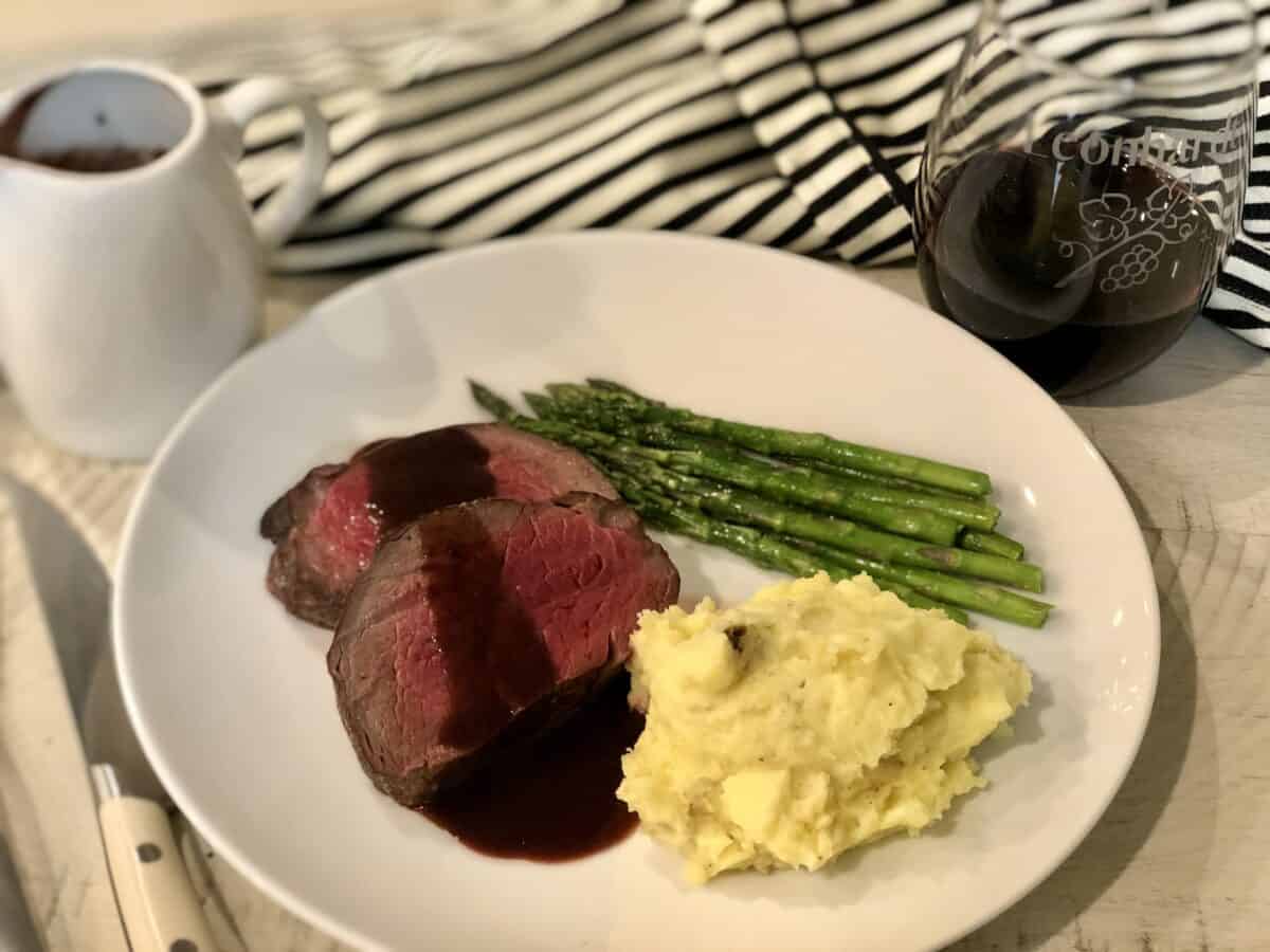 a white plate with medium cooked beef tenderloin served with red wine sauce mashed potatoes and asparagus a glass of red wine and a serving dish of sauce with a black and white apron in the backround
