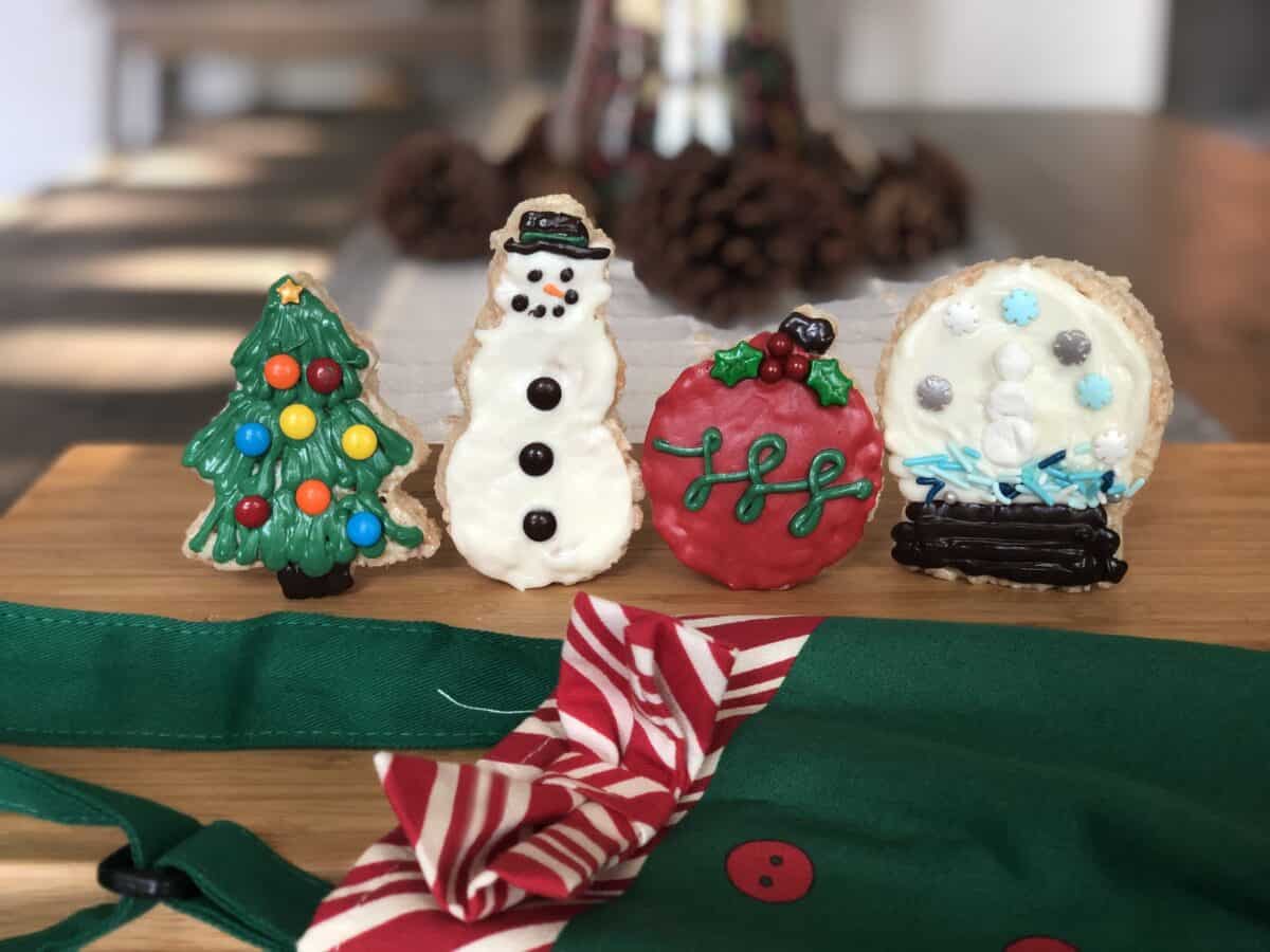 4 decorated christmas rice krispie treats in the shape of a christmas tree snowman ornament and snow globe are standing up on a brown cutting board with a green and red apron showing and pine cone in the background