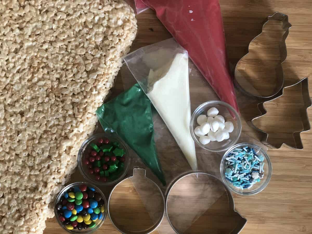a large sheet of rice kristie treat on a wood cutting board with bags of white green and red chocolate next to cookie cutters marshmallows mini m&ms and christmas sprinkles