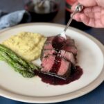 sliced steak spoon pouring over red wine sauce mashed potatoes asparagus on white plate