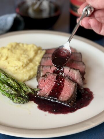 sliced steak spoon pouring over red wine sauce mashed potatoes asparagus on white plate