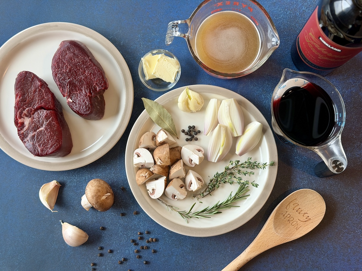2 steaks mushrooms garlic herbs shallots red wine and beef stock measured wood spoon over blue background 