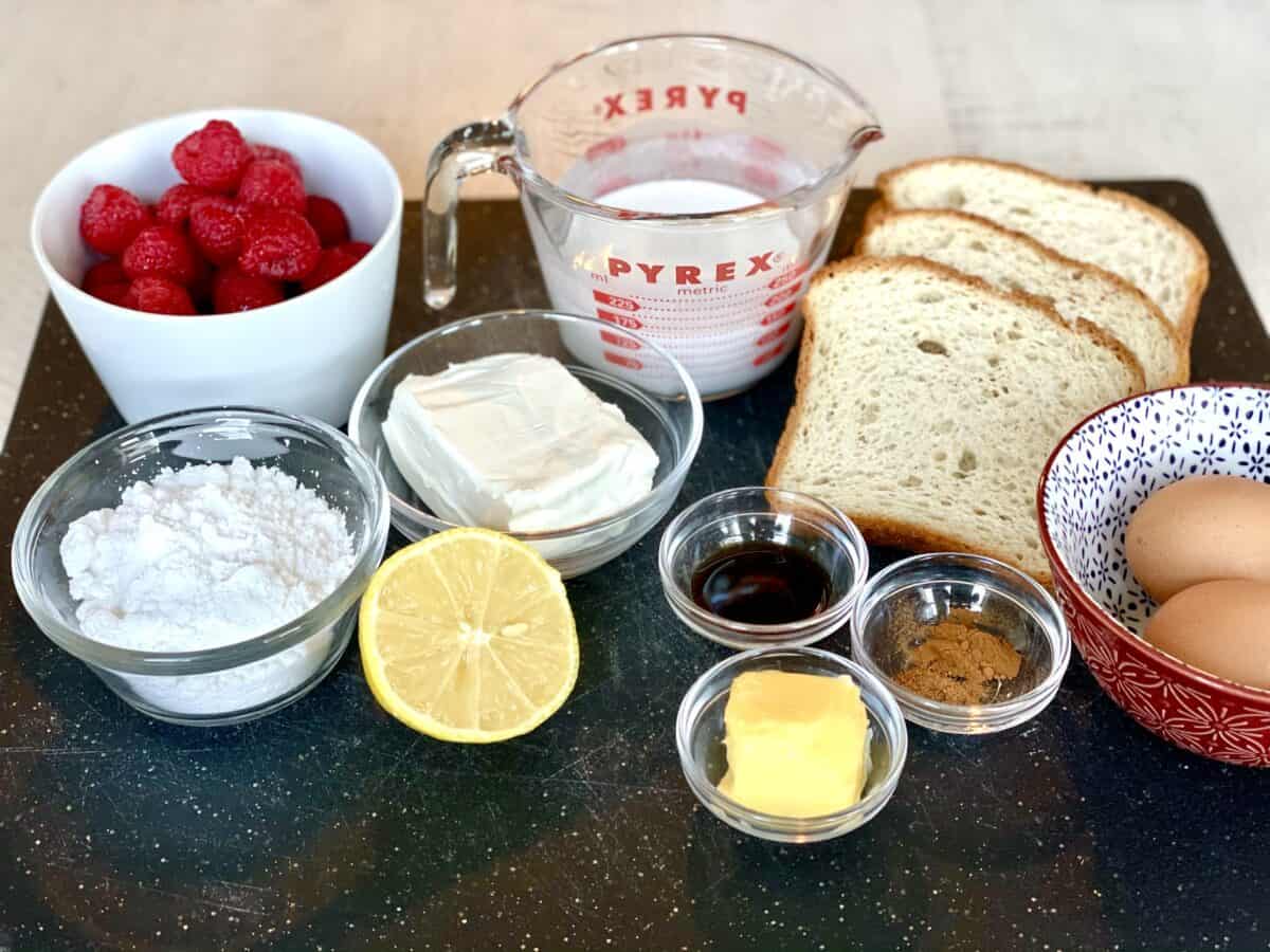 a white bowl of raspberries a glass bowl of powdered sugar half a lemon half a block of cream cheese half a cup pf half and half 4 slices of bread a small bowl of vanilla extract a small bowl of butter a small bowl of spices and 2 brown eggs in a red bowl