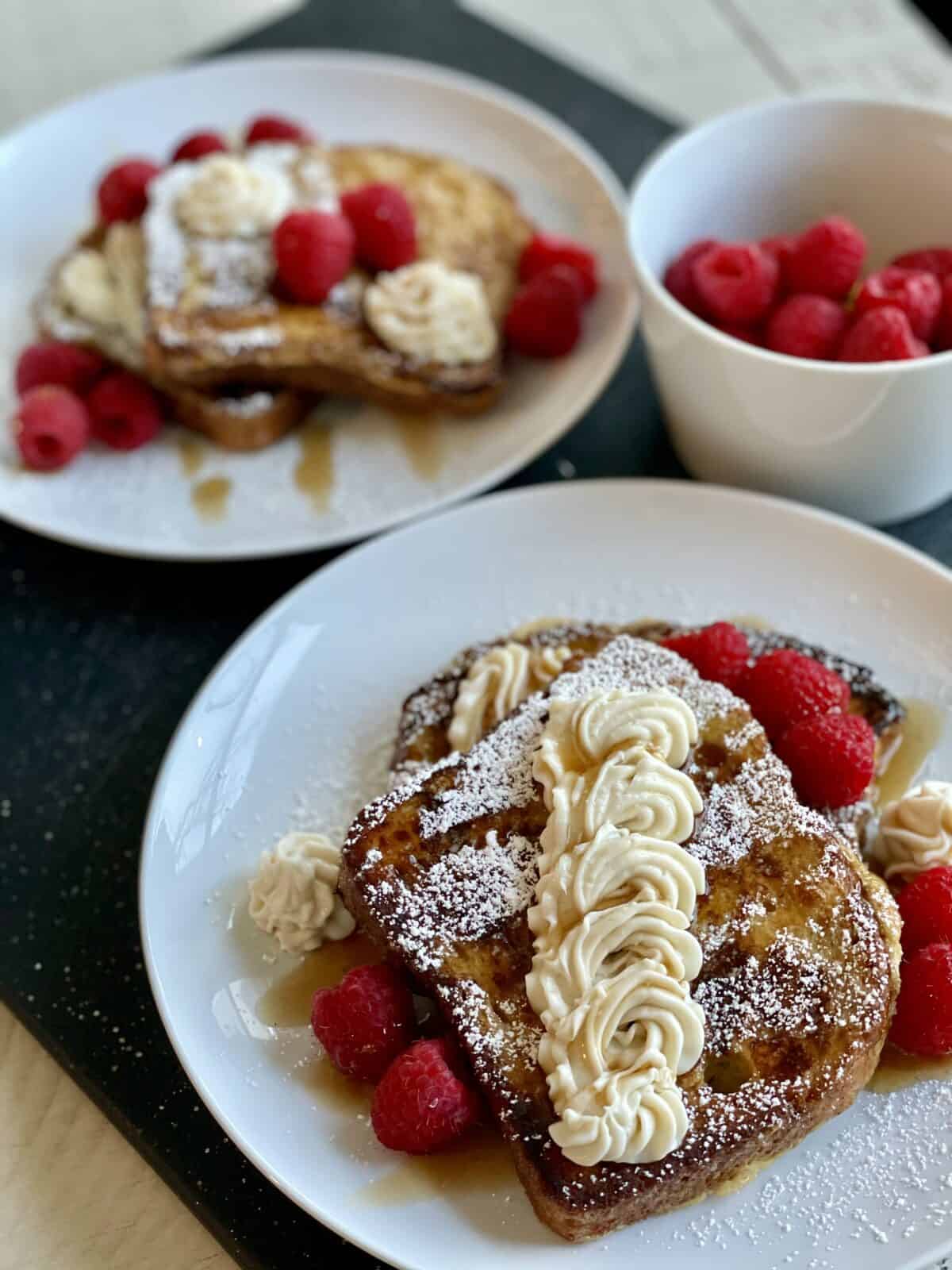 2 white plates with cooked french toast dressed up with cheesecake filling powdered sugar fresh raspberries and maple syrup