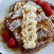 2 white plates with cooked french toast dressed up with cheesecake filling powdered sugar fresh raspberries and maple syrup