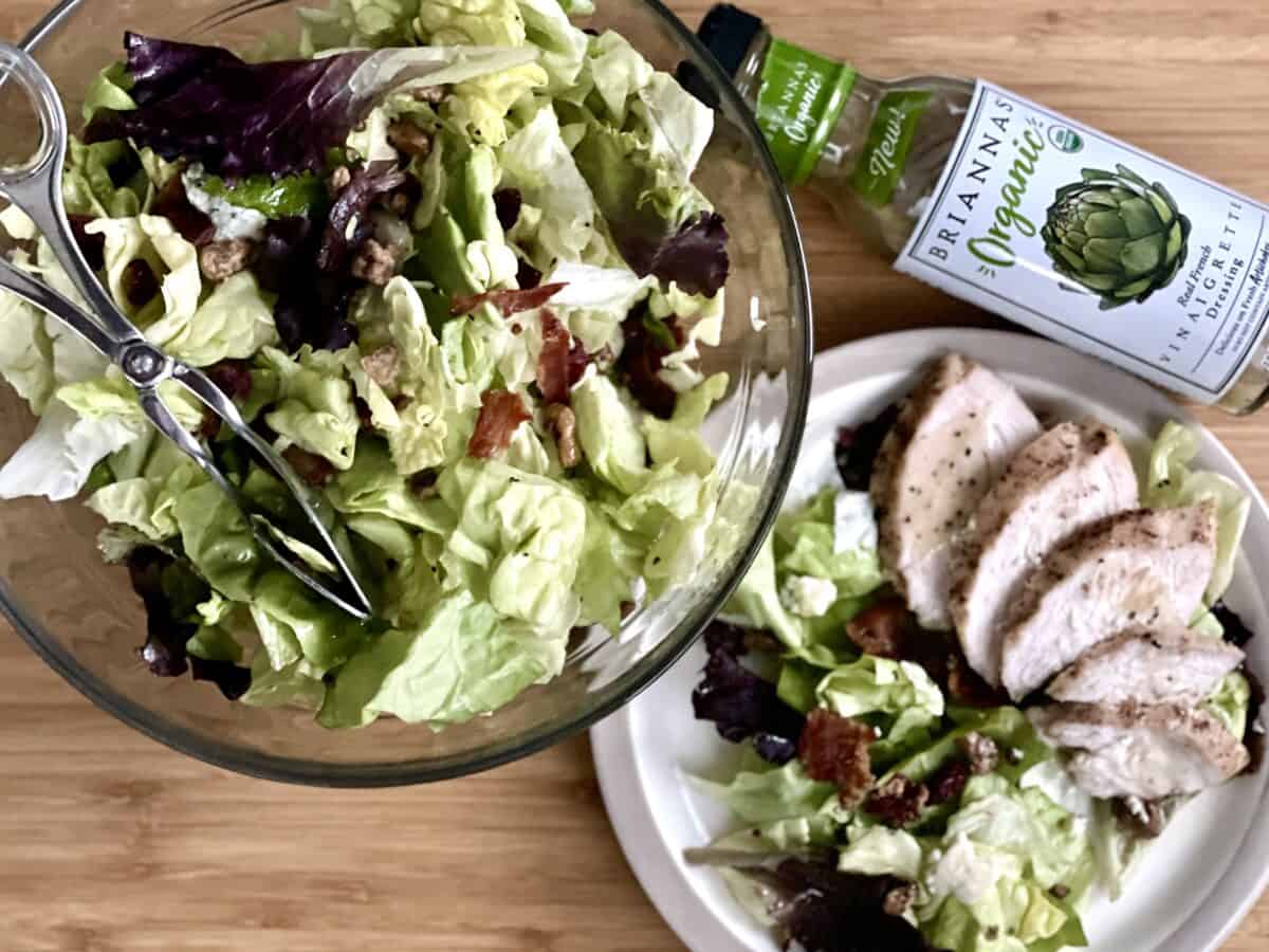 A light wood cutting board with a glass bowl filled with cranberry walnut and blue cheese salad next to a plate with salad and grilled chicken 