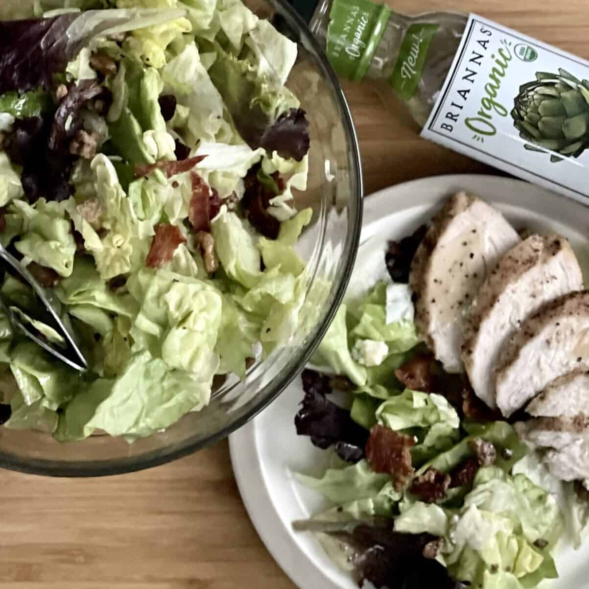 A light wood cutting board with a glass bowl filled with cranberry walnut and blue cheese salad next to a plate with salad and grilled chicken