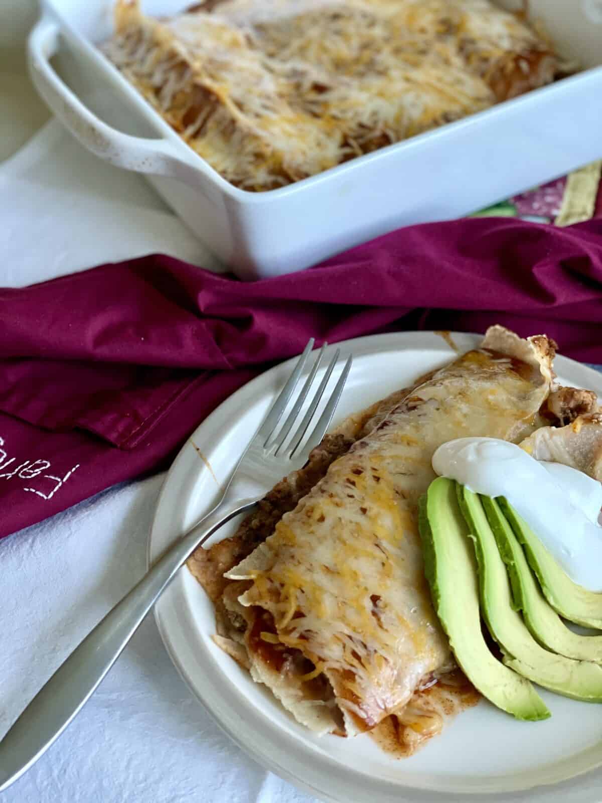 cooked beef and bean enchiritos on a white plate with sliced avocado and sour cream with the baking dish and more enchiritos behind the plate