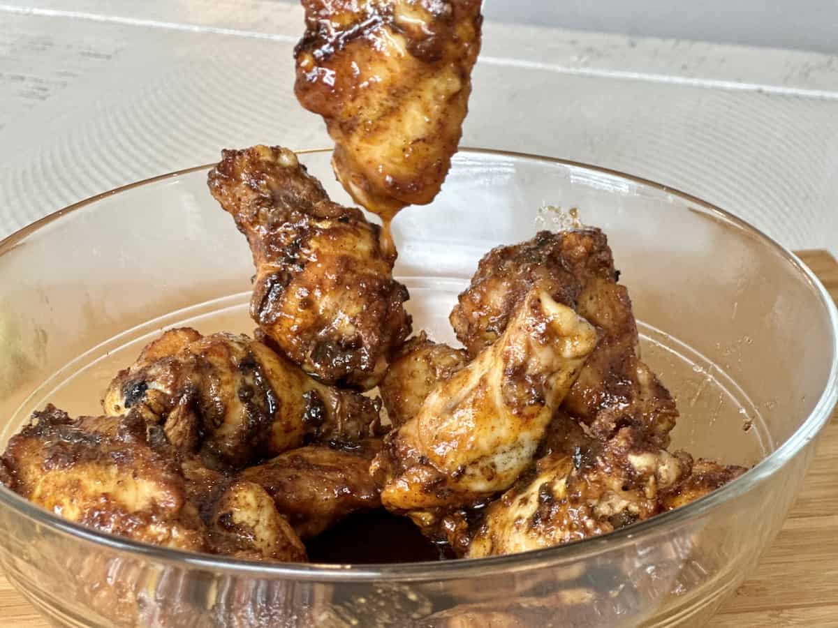 saucy chicken wing being held up over a bowl of cooked chicken wings
