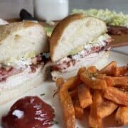 a white plate with a chicken cob sandwich cut on a diagonal with sweet potato fries ketchup and a jar of blue cheesing in the background