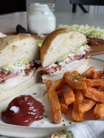 a white plate with a chicken cob sandwich cut on a diagonal with sweet potato fries ketchup and a jar of blue cheesing in the background 