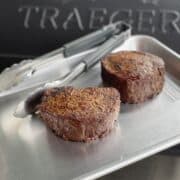 two perfectly cooked prime filets on a quarter sheet pan with tongs and a traeger smoker in the background