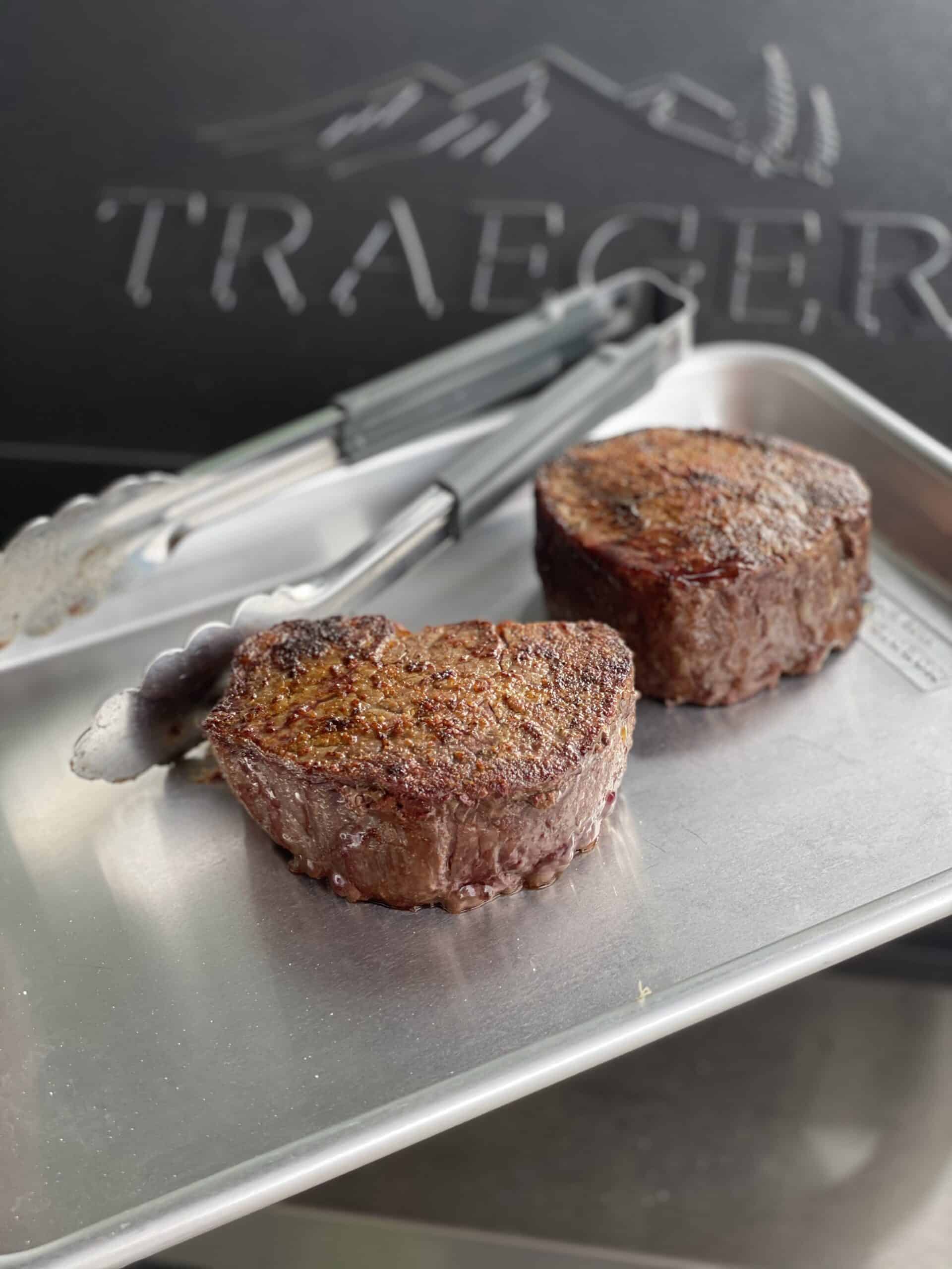 two perfectly cooked prime filets on a quarter sheet pan with tongs and a traeger smoker in the background