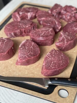 a brown cutting board with 11 trimmed filet mignon and a trimming knife