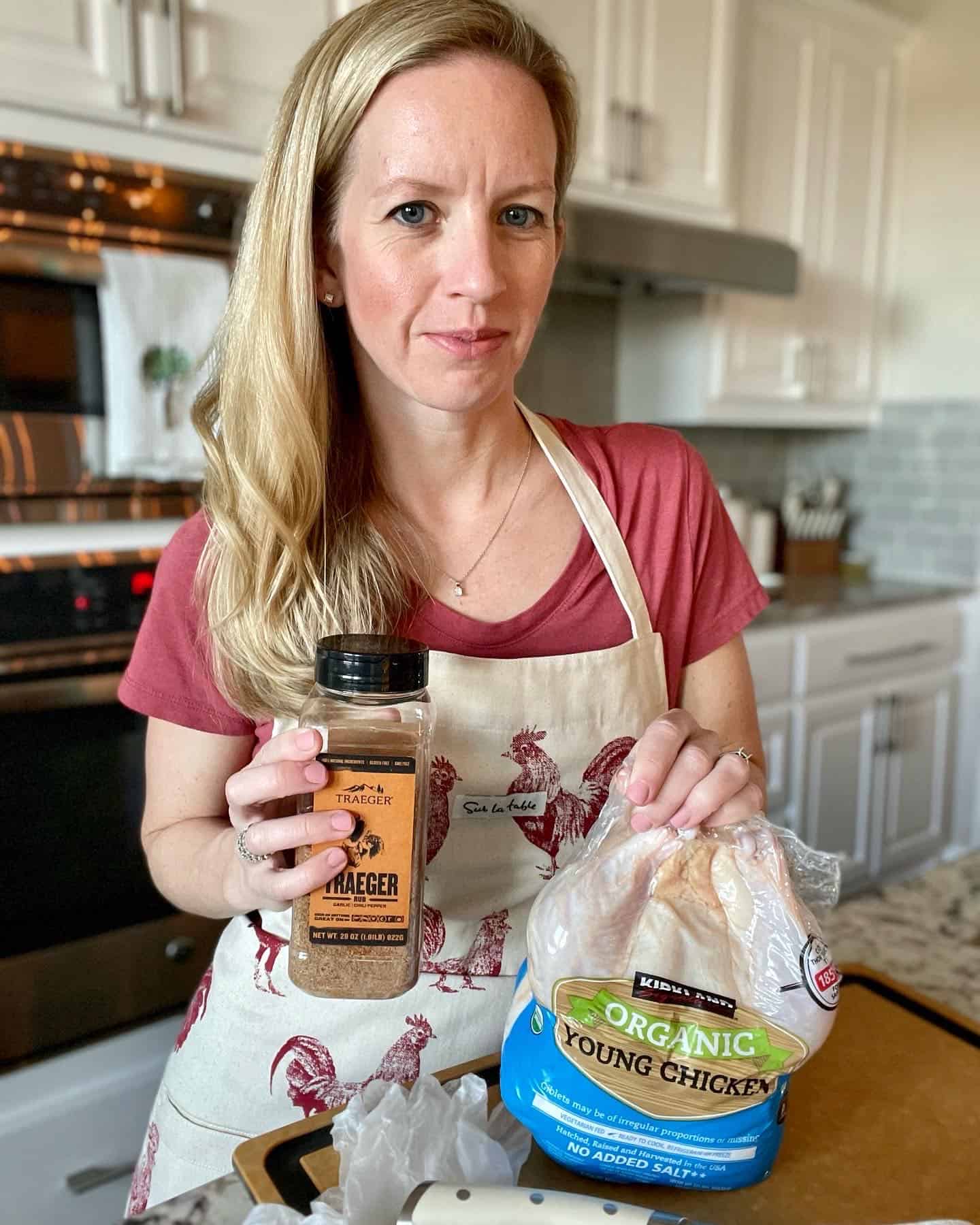 alexis wearing an apron with chickens on it and  holding a packaged whole organic chicken and a bottle of traeger spice rub 