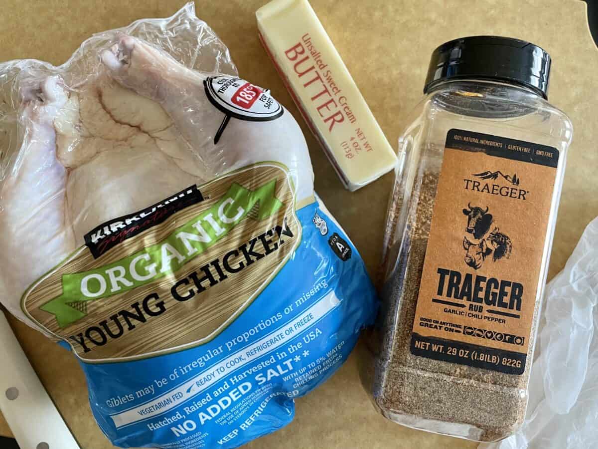 a whole raw chicken in its plastic packaging on a cutting board next to a stick of butter and bottle of traeger rub