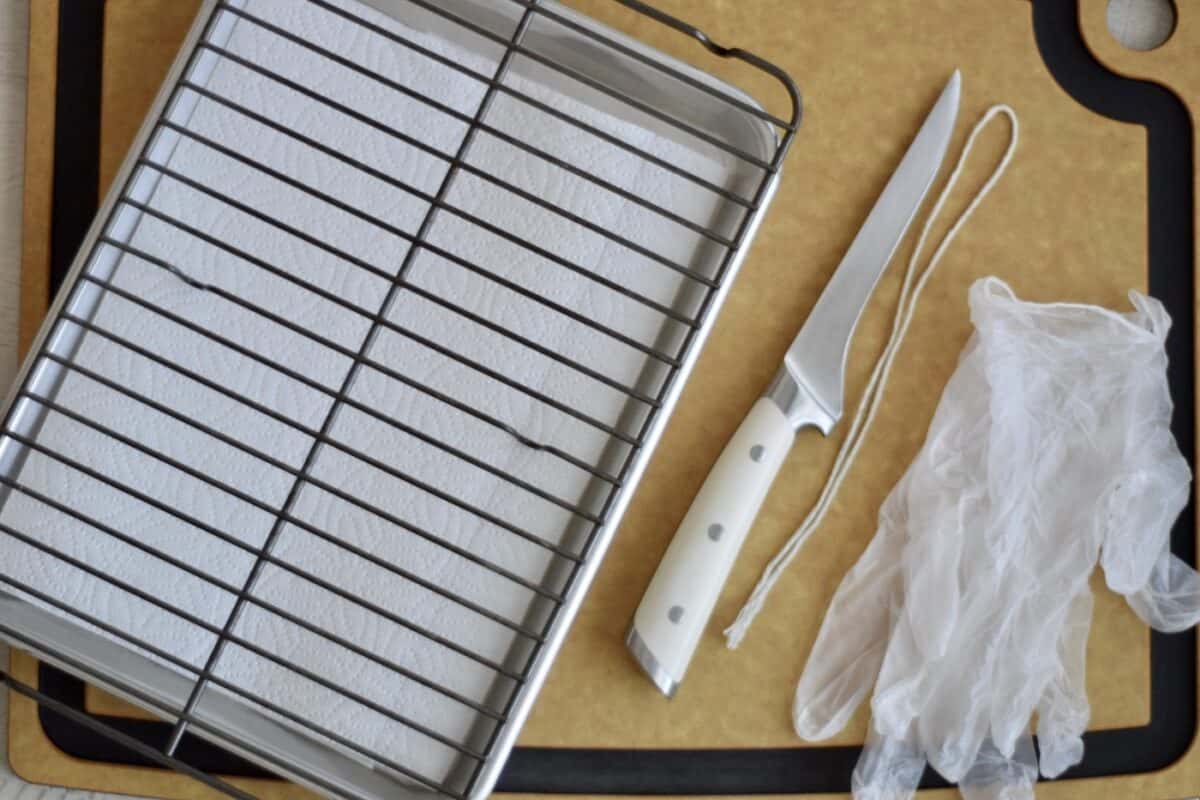 a brown cutting with a sheet pan lined with a paper towel and wire rack on top next to a sharp boning knife kitchen string and food safe gloves