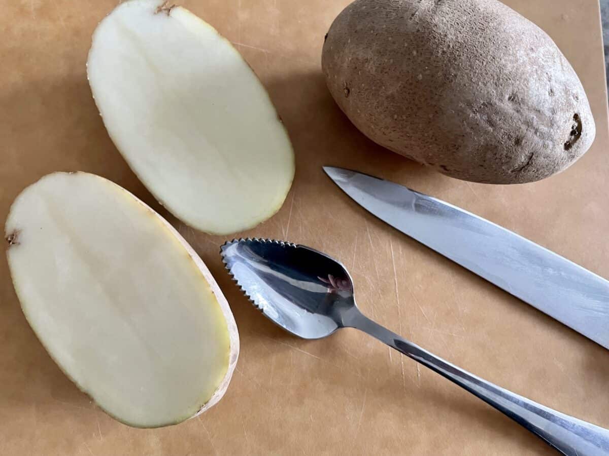 one russet potato cut in half and one whole russet potato on a cutting board with a sharp knife and grapefruit spoon