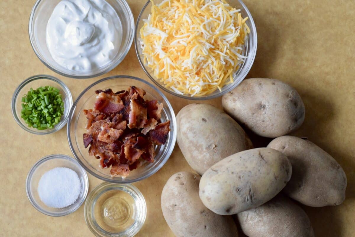 a brown cutting board with potato skin ingredients including small russet potatoes a bowl of shredded cheddar cheese a bowl of crispy bacon a small bowl of sour cream very small bowl of chopped chives sea salt and vegetable oil
