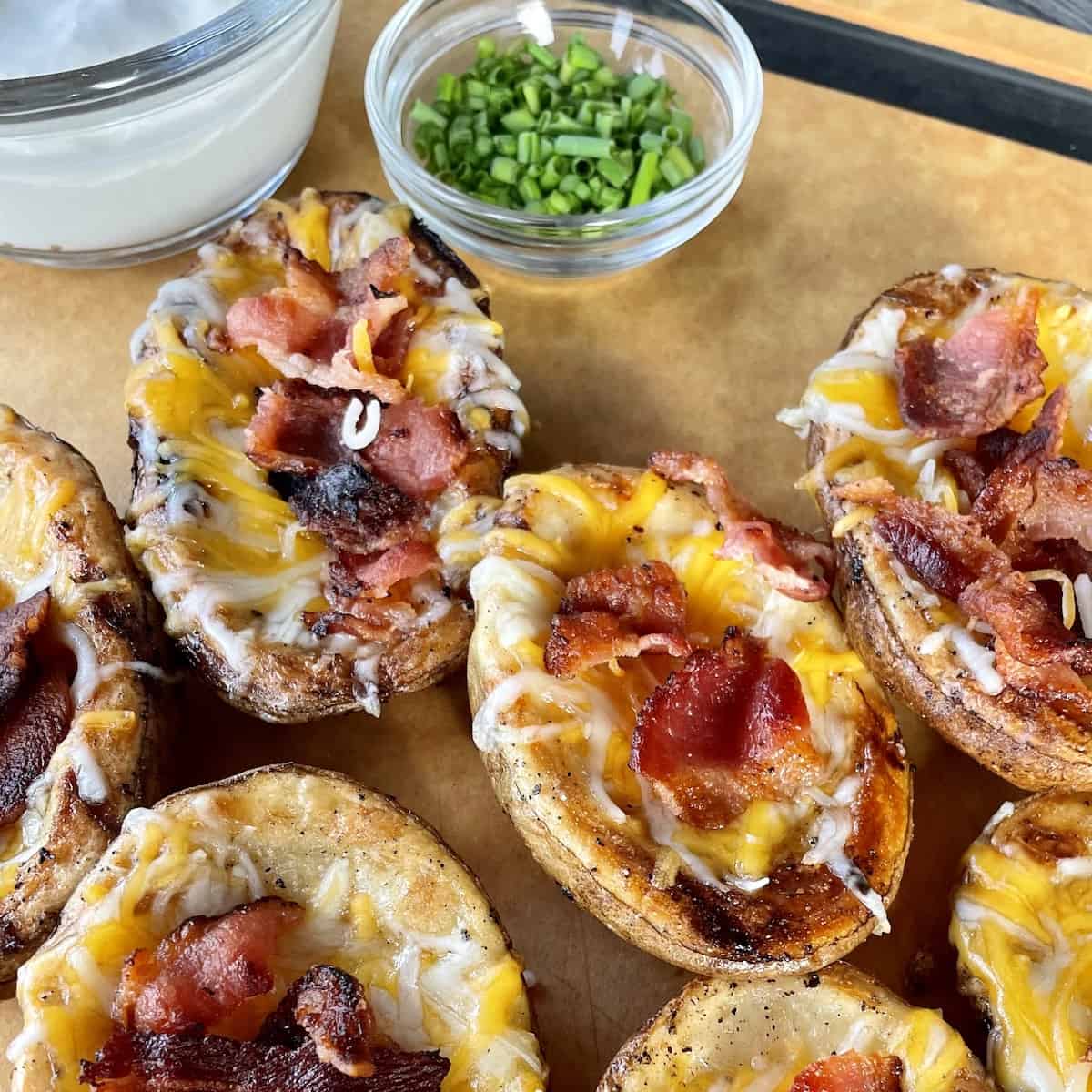 brown cutting board with 6 cooked potato skins filled with melted cheese and crispy bacon with a cup pf sour cream and bowl of chopped chives for topping