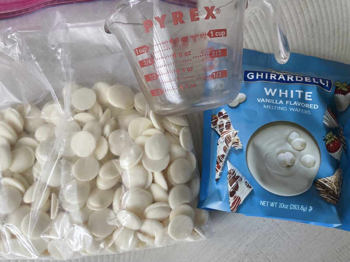 a large zip top bag of merkens white chocolate melting wafers next to glass measuring cup and ghirardehli melting wafers on a white kitchen table