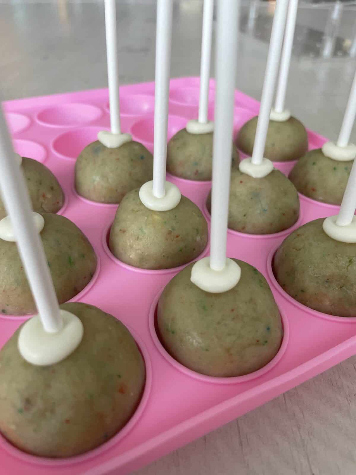 12 cake pops being held up in a pink silicone mold with the lollipop sticks in them ready for dipping